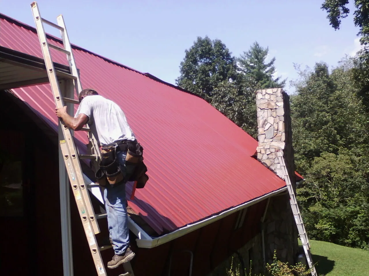 Roofer on a ladder beside a red metal roof and stone chimney — Soffit & Fascia Repair by Roofing Brothers in Newport
